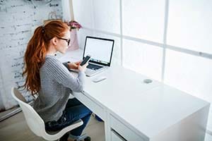 Red hair woman working at computer with phone in hand.
